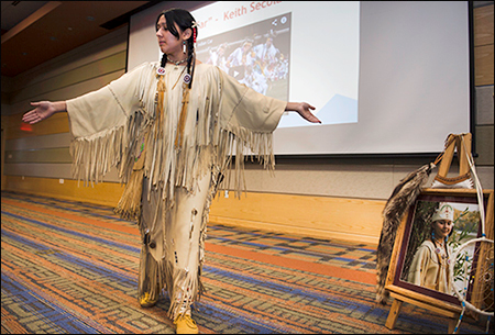 This is a photo from a previous Native American Heritage Month Celebration. The presenter is wearing their traditional attire with braided hair. They are standing with their left leg in front of their right leg and their arms extended out. 
