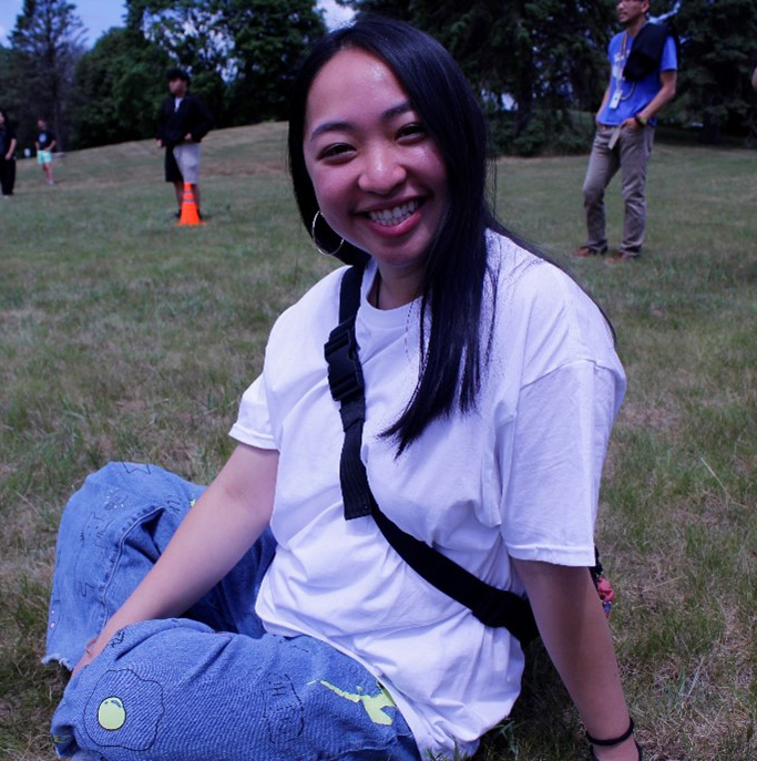 Photo of Mai Nou, a female with long black hair wearing a white tshirt and jeans with a black bag around her shoulder. She is sitting in the grass with people standing in the distance.