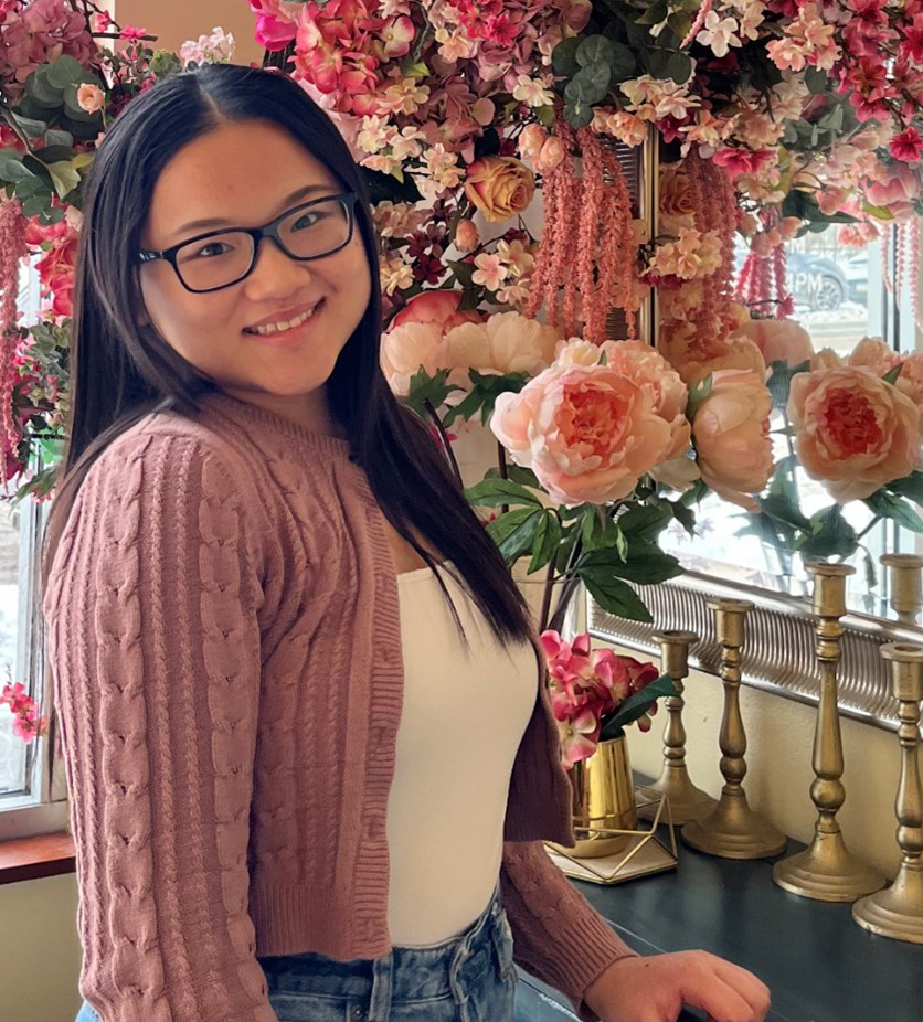 Photo of Jazlynn Vang, an Asian woman wearing a pink cropped sweater, white tank top and blue jeans. She has long black hair and is wearing black glasses and is standing in front of a large floral arrangement made up of white and pink roses and peonies.