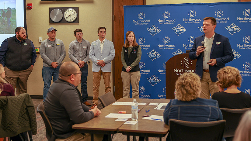 A man wearing a blue suit jacket and khakis holds a microphone and speaks to a group of people sitting at tables. He's joined by five people standing to his right and all are standing in front of a blue backdrop that says Northwoods Technical College.