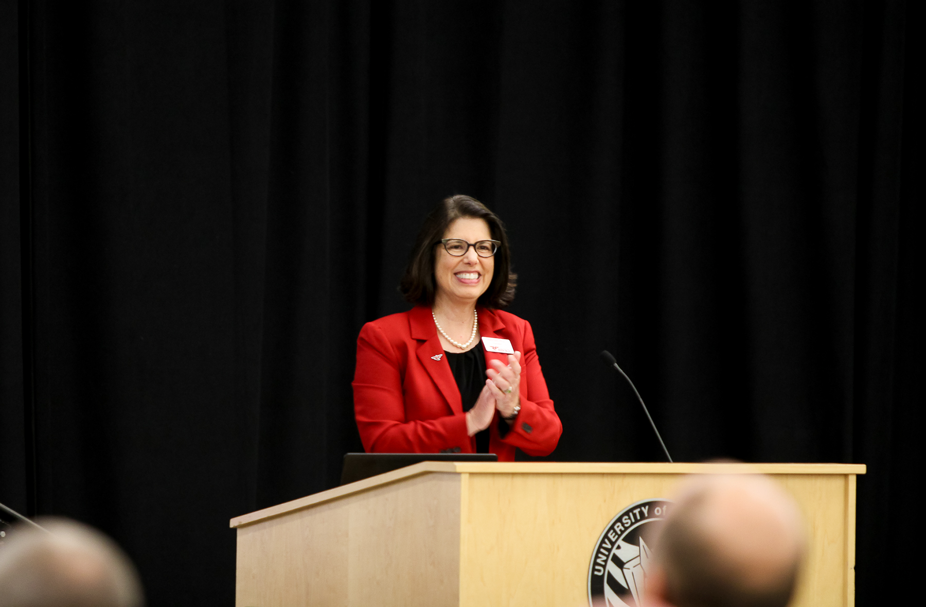 Chancellor Gallo, wearing a a red suit jacket, black shirt, pearl necklace and black glasses, claps while standing at a podium and addressing the crowd at Opening Meeting in August 2023