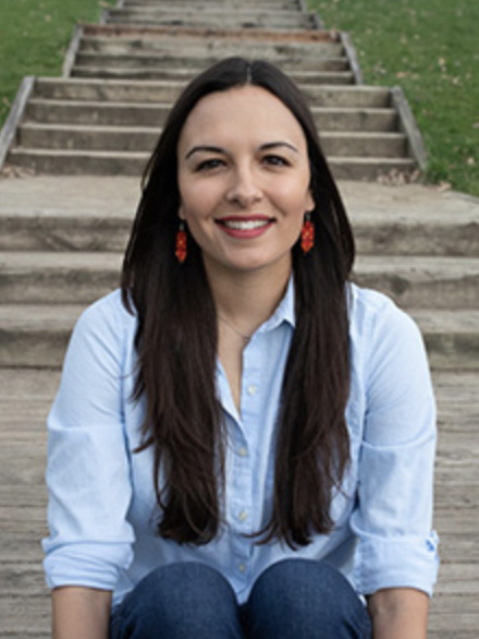 A headshot of Katrina Phillips, a woman looking directly at the camera with long dark hair, a blue button down shirt and red earrings. She is seated on an outdoor staircase. 
