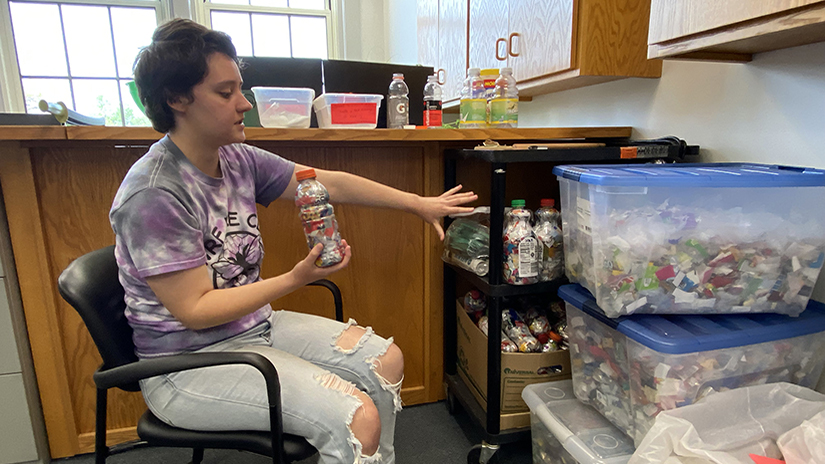 Amber Rappl wears a purple tshirt and sits in a chair holding a plastic bottle. Next to her are several totes filled with recycled plastic bottles and wrappers.