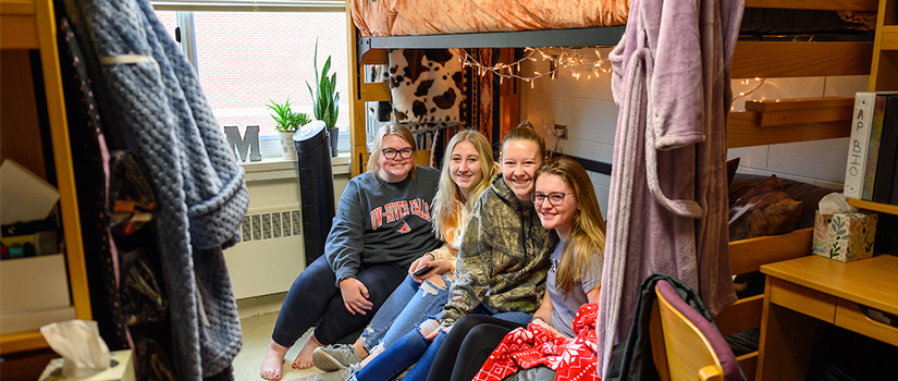 Four students smile together while hanging out in a Hathorn Hall room under a lofted bed.
