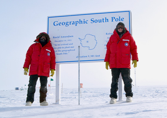 Two male students stand in front of a sign at the geographic south pole in Antarctica