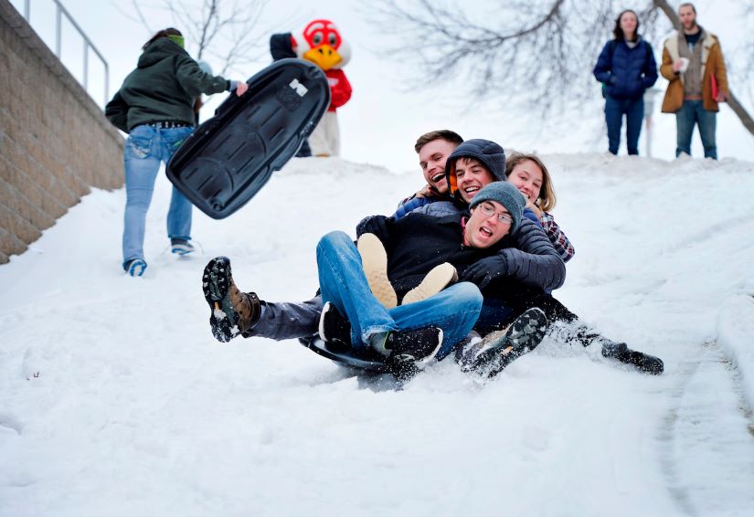 Four students hang onto each other as they are piled onto a sled heading down a hill in the winter.