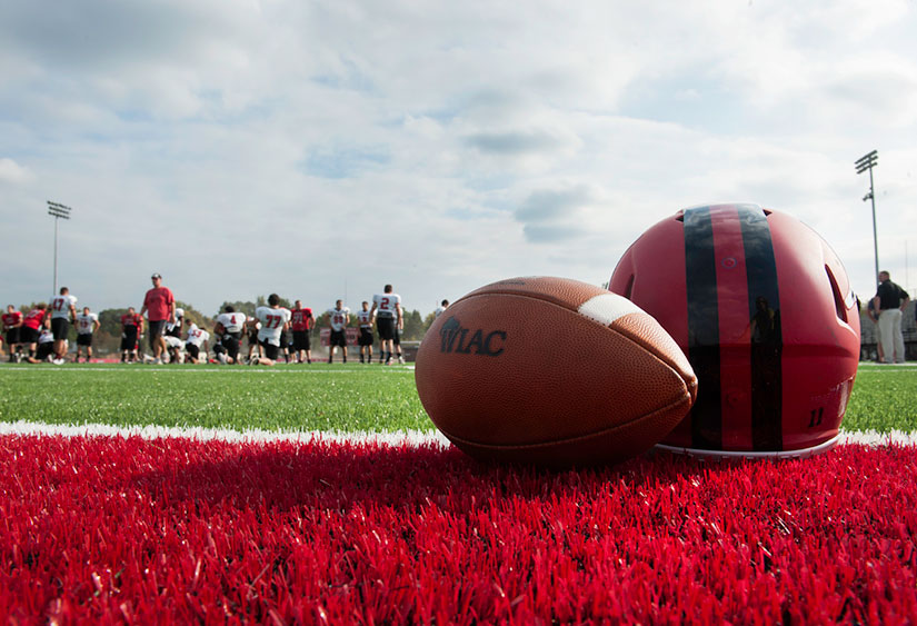 image of football and football helmet sitting on the new turf with football players practicing in the background.