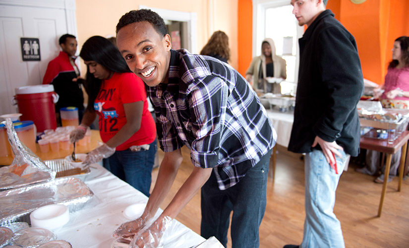 This is a photo from a past Soul Food Dinner, an event put on by BSU each year. The photo focuses on one student as he wears food safety plastic gloves, a black and white plaid shirt, jeans, and is smiling at the camera.
