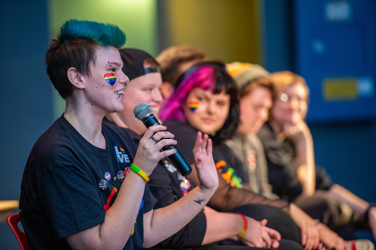 Person with blue hair and rainbow heart sticker on cheek speaks into a microphone while several other students look on