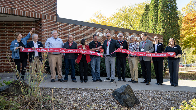 A line of UWRF administrators and donors stand in front of the Agriculture Science Building holing a red banner that reads Wuethrich/Grassland Family Dairy Center of Excellence. 