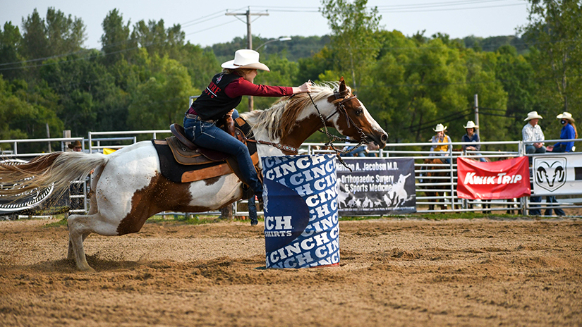 A rider wearing blue jeans, a red long sleeve shirt and black vest and white cowboy hat steers her brown and white paint horse around a barrel during a barrel racing event.