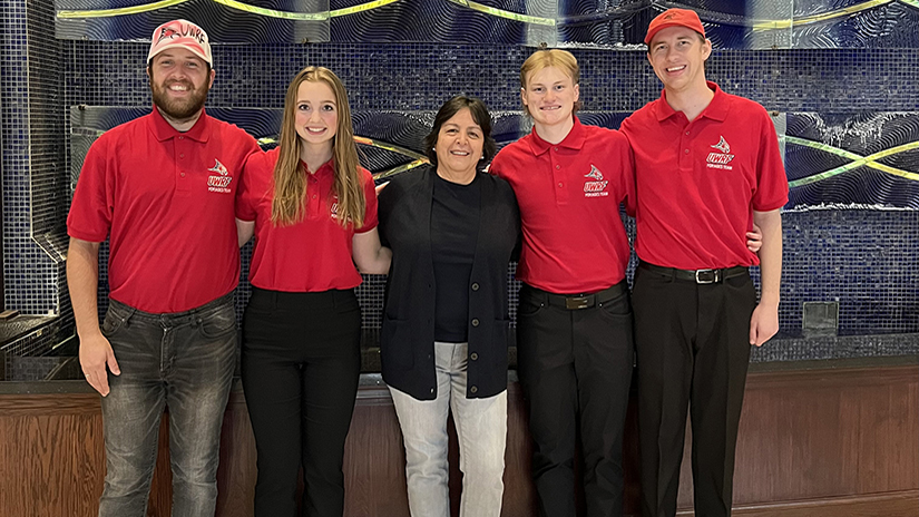 Four students, three males, one female, all wear red polos and dark pants pose with a faculty member wearing a dark shirt, sweater and jeans. There are two students on each side and the faculty member is in the middle. They are all posing and smiling.