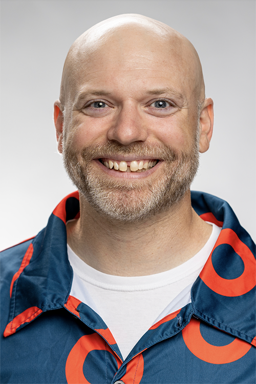 Headshot of Kevyn Juneau, smiling white male wearing a white tshirt and a navy blue button up shirt with orange circles on it