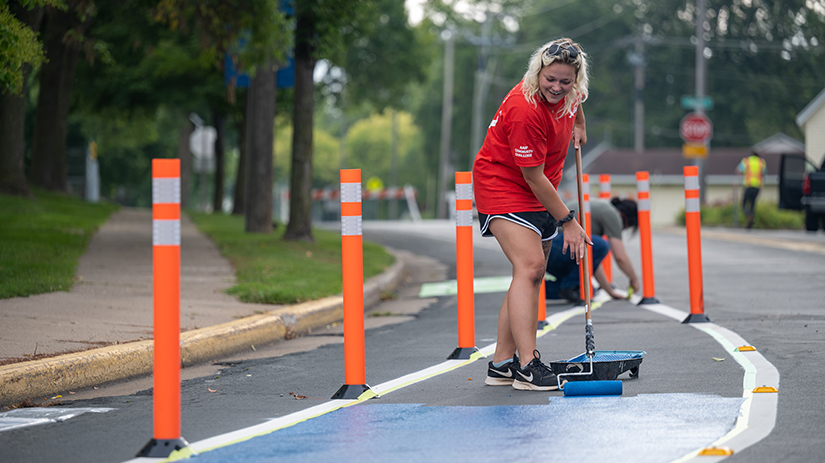 A blonde person wearing a red shirt and black shorts uses a rollerbrush to paint the street in River Falls