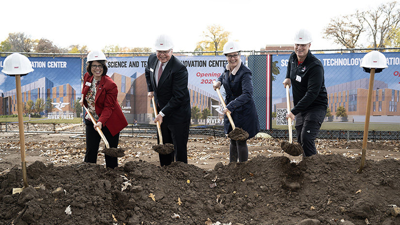 Chancellor Gallo, wearing red blazer, UW System President Jay Rothman, wearing dark suit, Kristi Cernohouse, wearing dark winter coat, and Jeff Cernohous, wearing dark sweater, all wearing white hard hats use gold shovels to dig up a piece of dirt