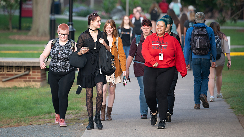 Large group of students walking on sidewalk through campus, laughing and talking with each other