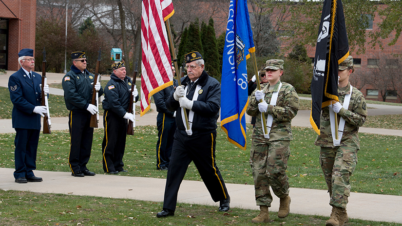 Participants take part in a past Veterans Day event at UW-River Falls. Three people in military uniforms carry American, Wisconsin and military flags while fellow uniformed people stand at attention