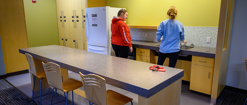 Ames pod kitchenette including island, seating, countertops, refrigerator, lockers, a microwave oven, and two students working together.