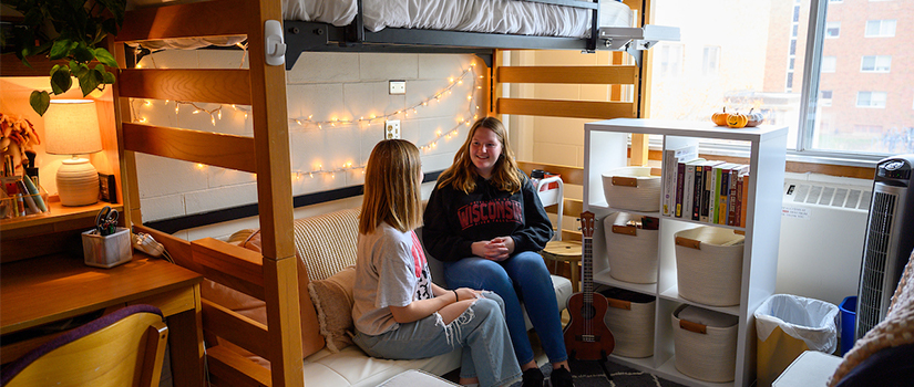 Two roommates talk to each other under a lofted bed.