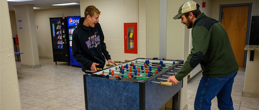 Two students play foosball in the McMillan basement. A beverage and snack machine are in the background.