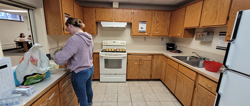 A student begins a cooking project in the basement kitchen of Grimm hall. The kitchen includes a refrigerator, oven, stove, microwave, sink, and counterspace