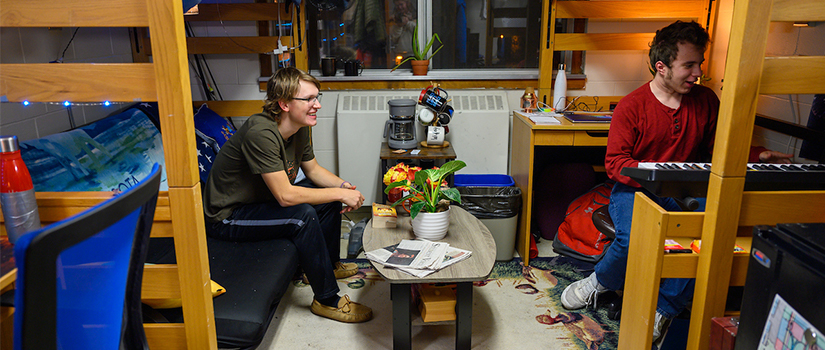 Two students sit under their lofted beds in the room to talk.