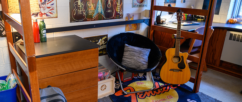 Inside a Stratton room, with a view of storage under a lofted bed. A dresser, chair, and guitar sit under the bed.