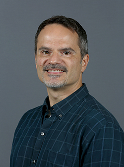 Keith Wojciechowski headshot. Male with grey and black hair, grey facial hair and a blue button down shirt