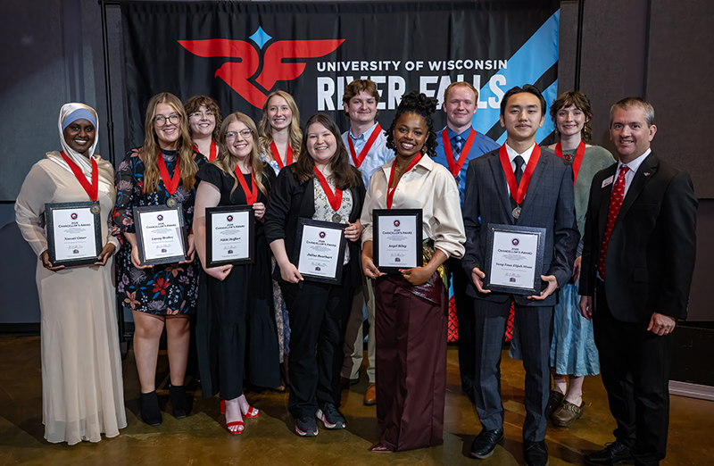two rows of people pose for a photo. All individuals but one are holding plaques and wearing red ribbons with gold medallions around their neck. They're all dressed in business casual clothing. 