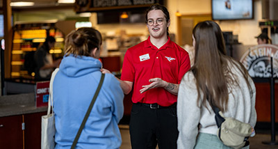 a person wearing a red polo and black dress pants speaks to two people who have their backs to the camera