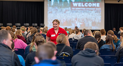 a female wearing a red polo shirt stands among chairs filled with people 