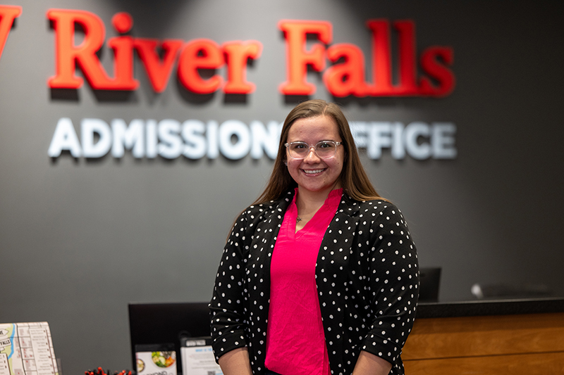 Afton Nelson stands in front of a desk and a sign that says "UW-River Falls Admissions Office"