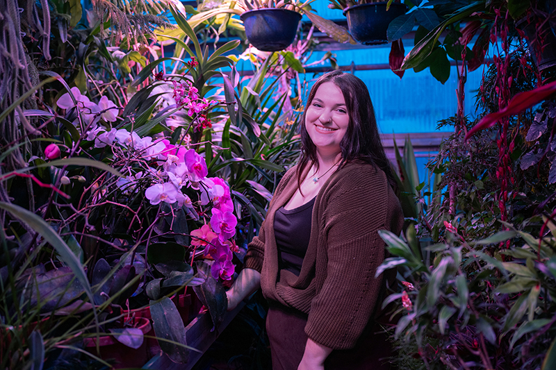 a white female stands in the middle of a greenhouse, surrounded by greenery