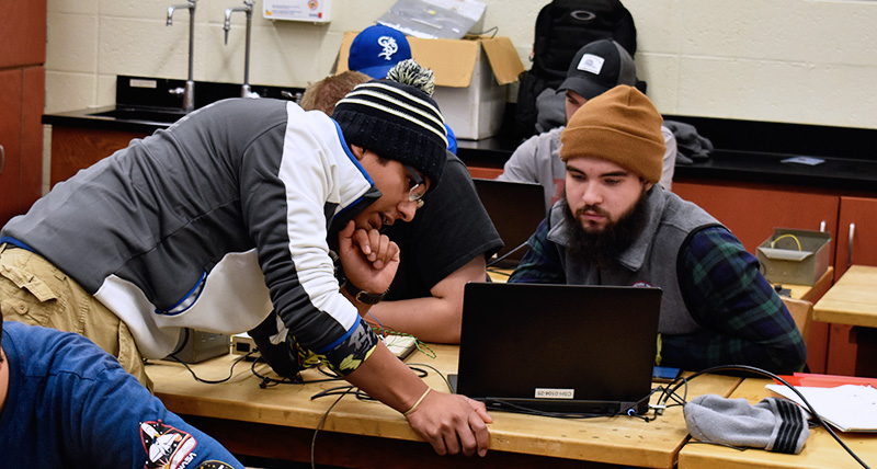 A student looks over another students desk to view their laptop screen in a physics lab classroom