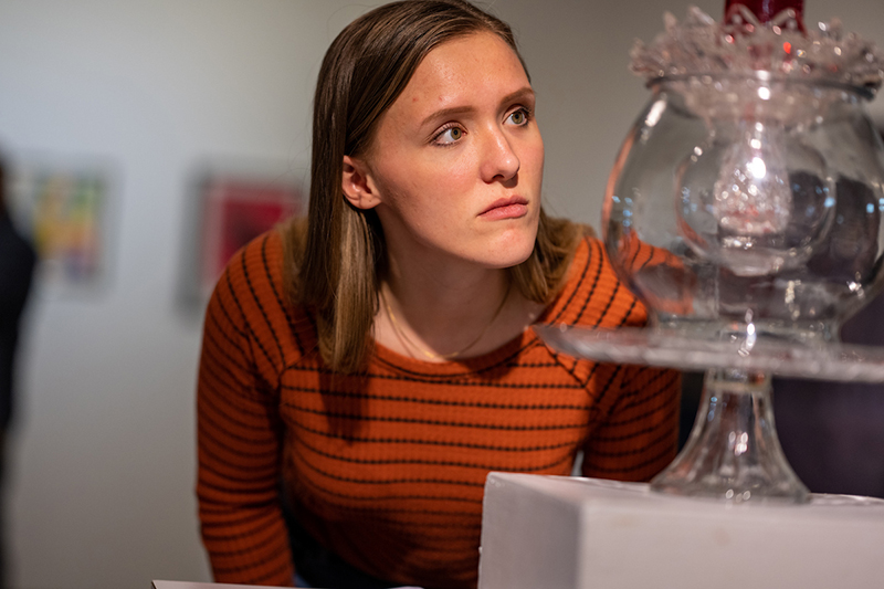 Woman with shoulder length brown hair wearing a long-sleeved orange shirt with black stripes looks closely at a glass artwork