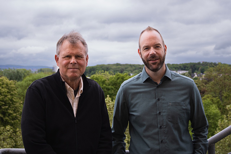 two men stand in front of a forested landscape. On the left is a man with short grey hair wearing a dark sweater. On the right is a man with very short dark hair and dark facial hair wearing a blue button up shirt.