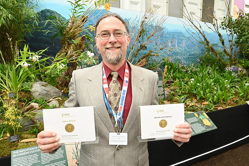 a man wearing a grey suit and red dress shirt holds up two award certificates.