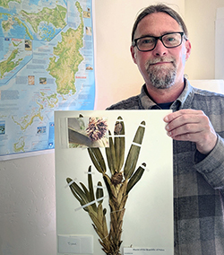 a man with glasses and gray facial hair holds a photo of an orchid plant 