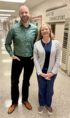 Two people stand together in a hallway. A taller man is on the left wearing a green button up shirt, black pants and brown shoes. A shorter woman with long grey/blonde hair stands on the right. She's wearing blue pants and a blue shirt and a white sweater.