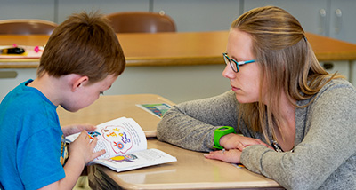 Student teacher kneels next to a child that is reading