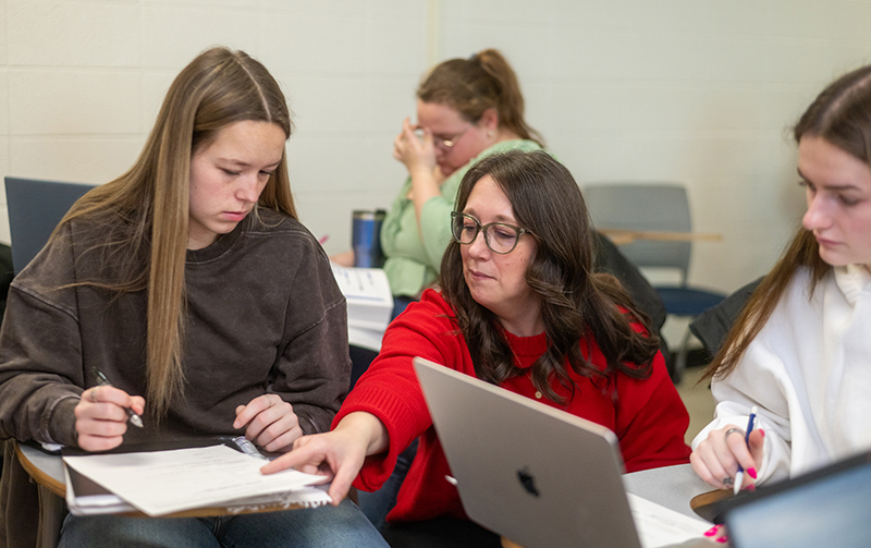 UW-River Falls Criminology ProfesDesiree Wiesen-Martin works with students in her Victimology class in February. Desiree is wearing a red sweater and has long brown hair and glasses. The two students both have long brown hair.