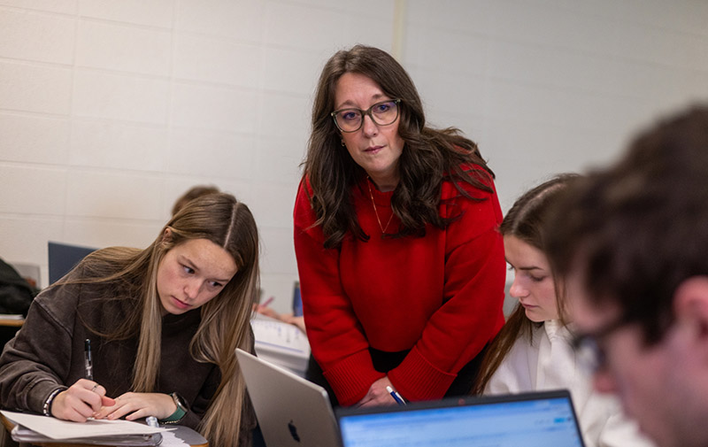 a professor with long brown hair and glasses is wearing a red sweater and is assisting a table full of students