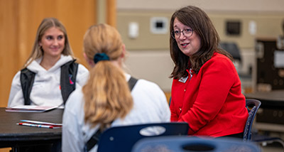 a brunette faculty member wearing a red top and glasses speaks with two high school female students while seated at a table