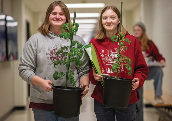 Two female students' side-by-side, carry potted plants down a hallway