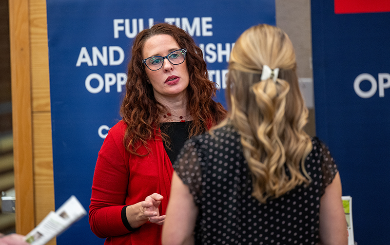 A woman with red hair wears a red sweater and glasses and speaks with a student at a Career Fair 