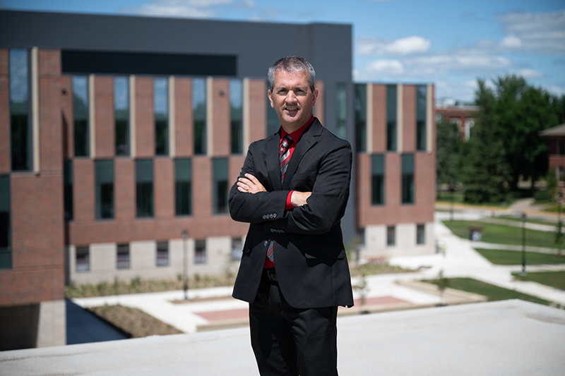 Chancellor John Chenoweth, wearing a black suit and red tie, stands with arms crossed in front of the SciTech building.