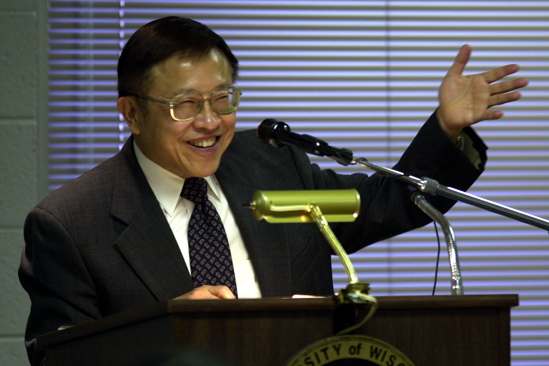 Man in a suit and tie gestures during a speech at a podium