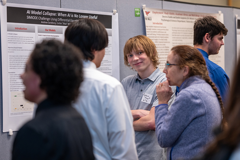 a person with shaggy blonde hair, a blue polo shirt and crossed arms stands in front of a posterboard while people looking at the poster stand around him