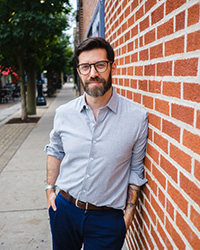 a man with dark hair and dark facial hair and glasses leans against a brick wall