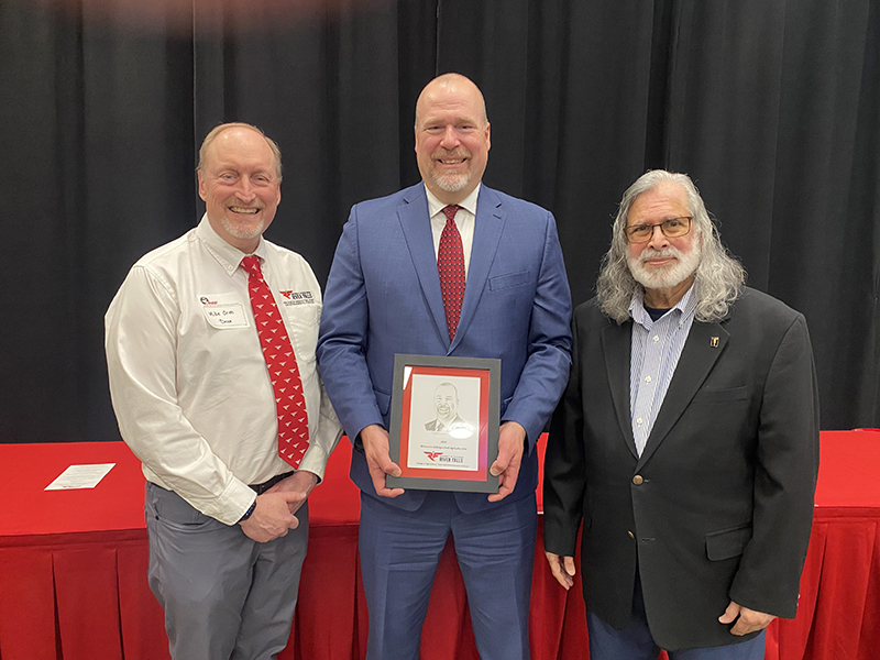 Three men pose for a photo. Man on left is wearing white dress shirt, red tie and dark grey dress pants. Man in middle is taller, wearing blue suit and red tie and holding a black and red plaque. Man on the right is shorter, has shoulder length grey hair and a beard and is wearing a dark sport coat and lighter dress shirt.
